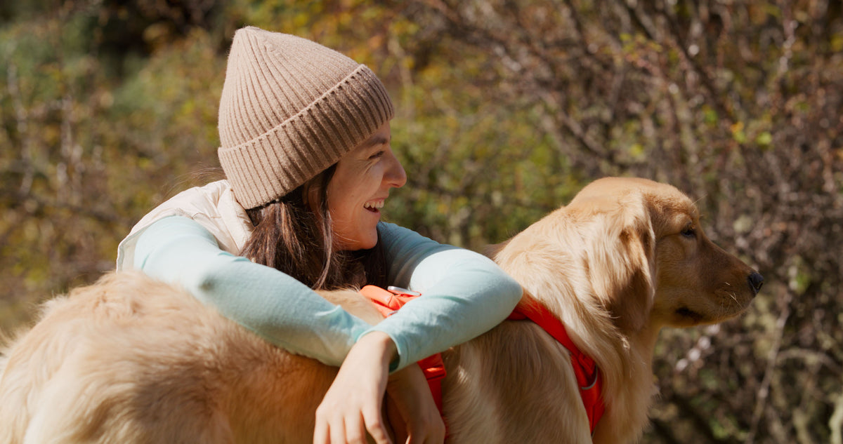 Woman in a beige knit hat and light blue jacket leaning on a dog's back with a blurred natural background