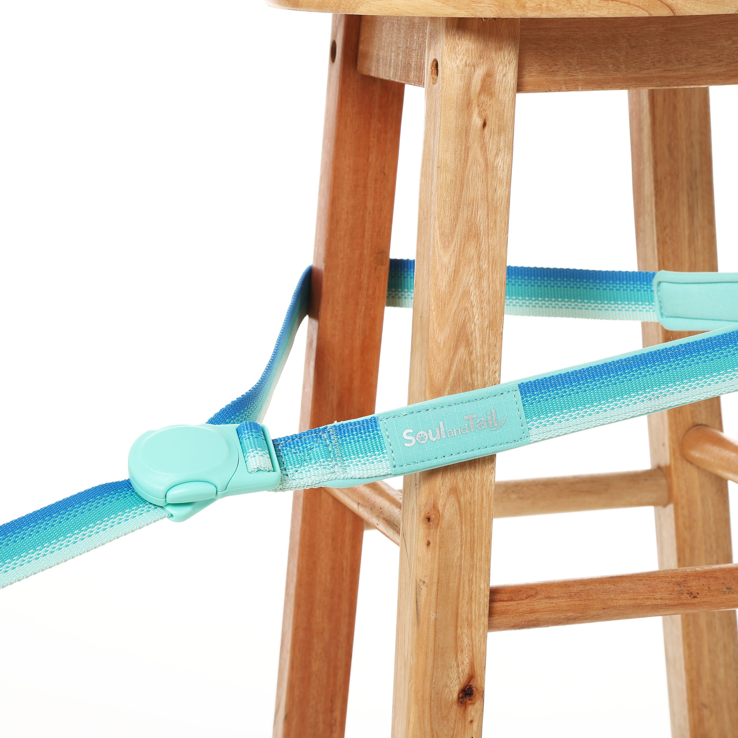 Turquoise dog leash with gradient design attached by a magnetic buckle to a wooden stool on a white background